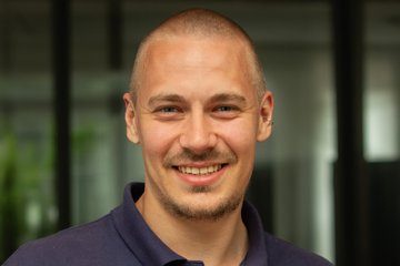 Eine Person in einem dunkelblauen Polohemd steht vor einem Gebäude mit großen Glasfenstern, Pflanzen im Hintergrund.