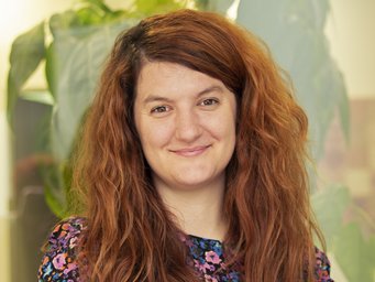 Freundlich lächelnd dreinblickende Frau mittleren Alters, wallenden rotblonden, zur rechten Kopfseite einem Scheitel folgenden dichten Haares steht vor einem Hintergrund, der eine überdimensionierte Darstellung einer großblättrigen Pflanze erkennen lässt. Sie vermittelt Anmut und Friedfertigkeit.