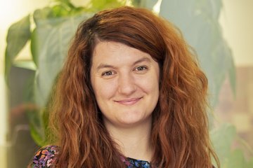 Freundlich lächelnd dreinblickende Frau mittleren Alters, wallenden rotblonden, zur rechten Kopfseite einem Scheitel folgenden dichten Haares steht vor einem Hintergrund, der eine überdimensionierte Darstellung einer großblättrigen Pflanze erkennen lässt. Sie vermittelt Anmut und Friedfertigkeit.