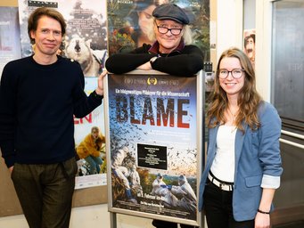 Drei Individuen markant unterschiedlicher Körpergröße stehen neben einem Plakat des Dokumentarfilms "Blame". Das Plakat enthält den Untertitel "Ein bildgewaltiges Plädoyer für die Wissenschaft" und zeigt Naturbilder. Die Individuen lächeln frohgelaunt in die Kamera der ablichtenden Person. Ganz offensichtlich sind sie von Freude und Stolz ergriffen.