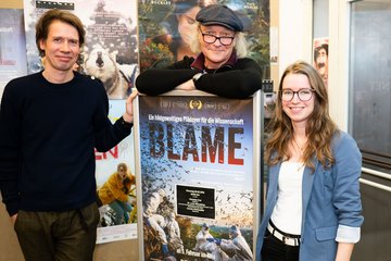 Drei Individuen markant unterschiedlicher Körpergröße stehen neben einem Plakat des Dokumentarfilms "Blame". Das Plakat enthält den Untertitel "Ein bildgewaltiges Plädoyer für die Wissenschaft" und zeigt Naturbilder. Die Individuen lächeln frohgelaunt in die Kamera der ablichtenden Person. Ganz offensichtlich sind sie von Freude und Stolz ergriffen.