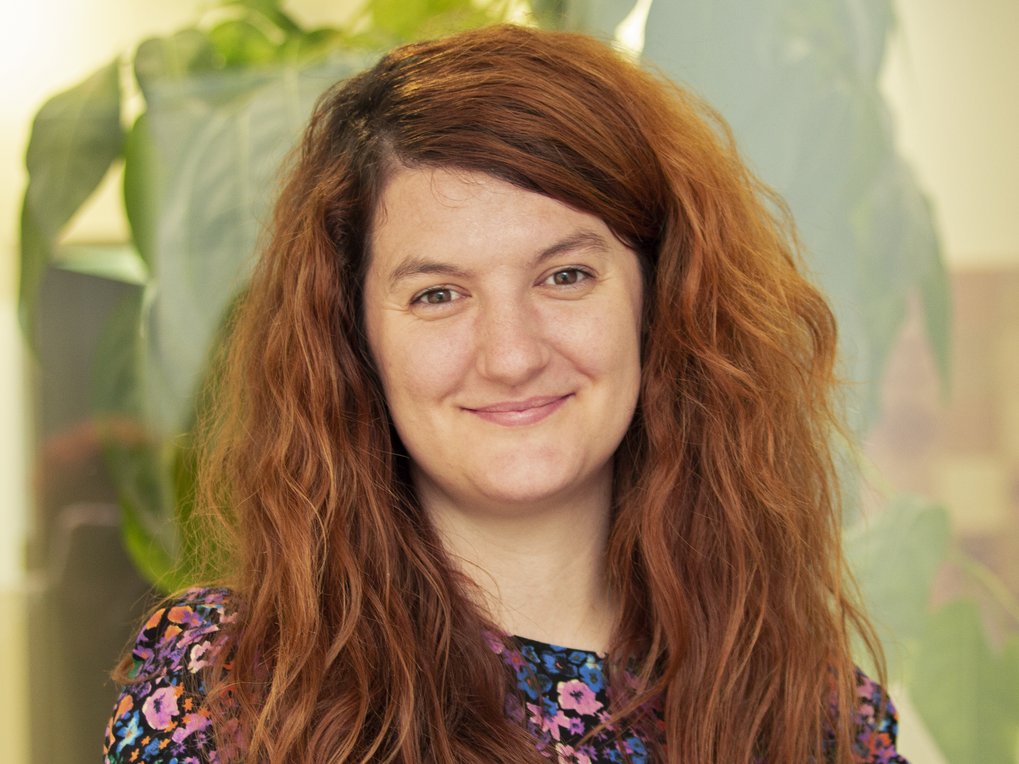 A middle-aged woman with a friendly smile and flowing reddish-blonde hair parted on the right side of her head stands in front of a background that reveals an oversized image of a large-leaved plant. She conveys grace and peacefulness.