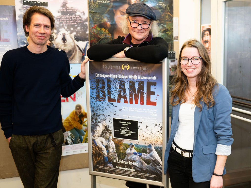 Three individuals of markedly different heights stand next to a poster for the documentary film “Blame.” The poster bears the subtitle “A visually stunning plea for science” and features images of nature. The individuals smile cheerfully at the camera. They are clearly overcome with joy and pride.