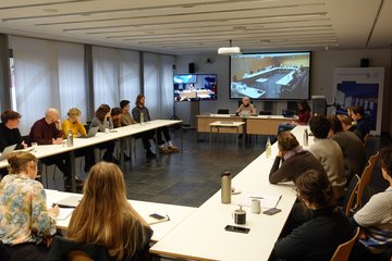 Eine Gruppe von Personen sitzt um einen langen Tisch in einem Konferenzraum herum und nimmt an einer Besprechung teil. Auf einem Bildschirm an der Vorderseite werden die Teilnehmer per Videokonferenz angezeigt. Auf dem Tisch stehen Laptops und Getränkeflaschen.
