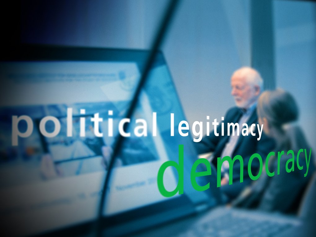 Overlay text "political legitimacy" and "democracy" on a laptop screen with people in a meeting room.