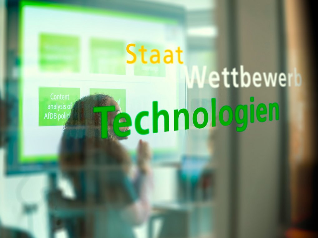 Begriffe "Staat", "Wettbewerb", "Technologien" erscheinen vor einem verschwommenen Präsentationsbild.