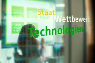 Begriffe "Staat", "Wettbewerb", "Technologien" erscheinen vor einem verschwommenen Präsentationsbild.
