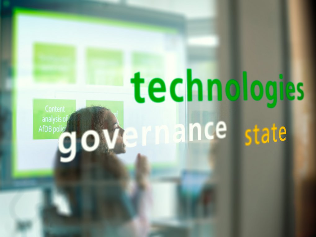 Words like "technologies," "governance," and "state" appear on glass, reflecting a screen's content.