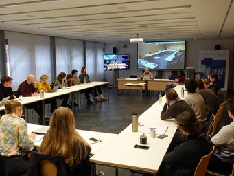 A group of people seated around a long table in a conference room, engaged in a meeting. A screen at the front displays remote attendees. Laptops and drink bottles are on the table.