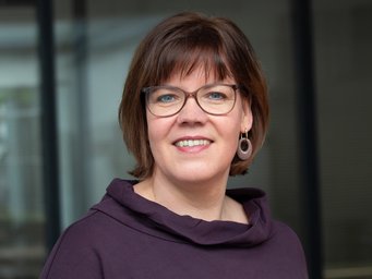 Eine Frau mit mittellangen braunen Haaren, bebrillt, trägt lila Oberteil und Ohrringe, steht vor einem unscharfen Hintergrund.