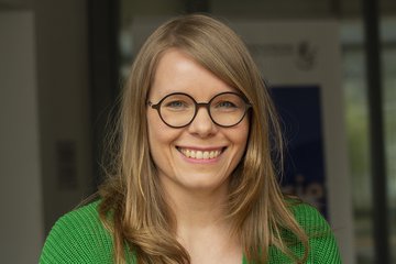 Eine Person mit langen, blonden Haaren trägt einen grünen Pullover. Das Gesicht ist verpixelt, Hintergrund unscharf mit einem Schild.