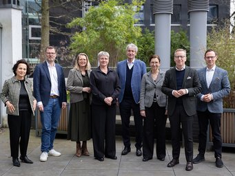 Acht Personen stehen auf einer Terrasse. Im Hintergrund sind hohe Pflanzen und moderne Gebäude zu sehen. Alle tragen formelle Kleidung.
