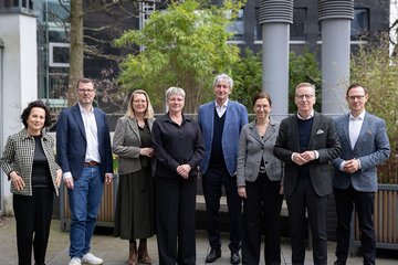 Acht Personen stehen auf einer Terrasse. Im Hintergrund sind hohe Pflanzen und moderne Gebäude zu sehen. Alle tragen formelle Kleidung.