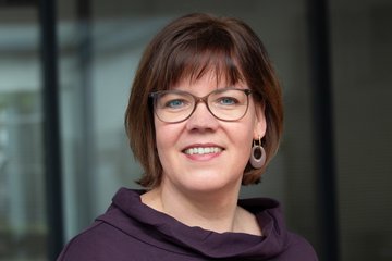 A woman with medium-length brown hair, wearing glasses, a purple top, and earrings, stands against a blurred background.