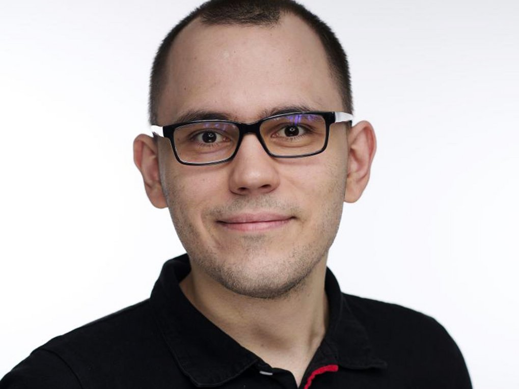 Portrait of a man with short hair wearing a black polo shirt against a neutral, light-colored background.