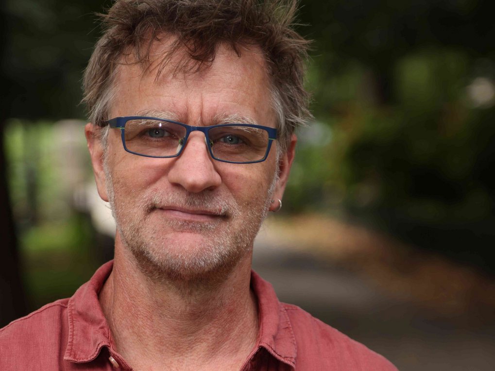 Portrait of a man in casual attire wearing a red shirt, with a blurred park path in the background.