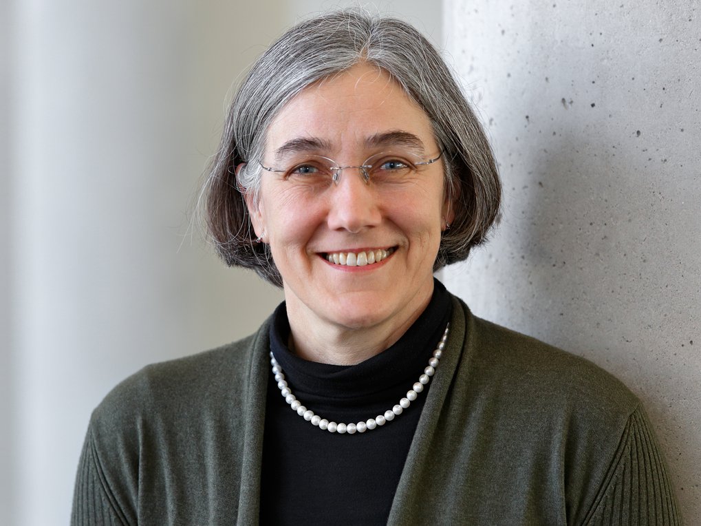 A woman with gray, chin-length hair is wearing a pearl necklace, a black turtleneck, and a green cardigan.