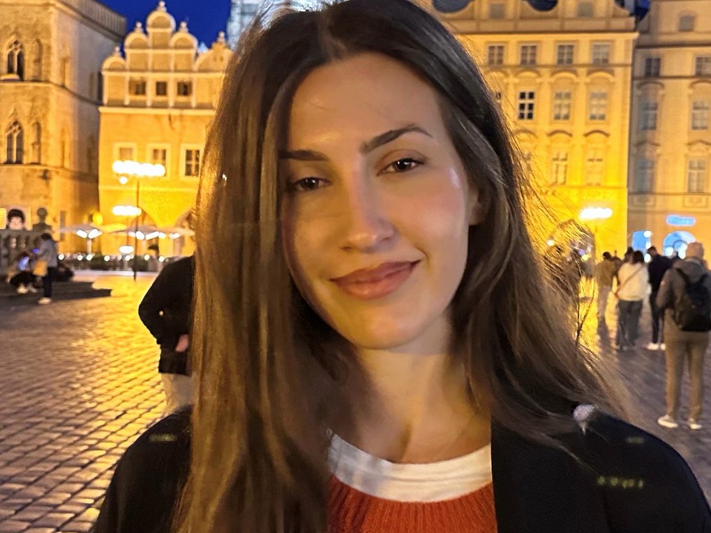A woman with long hair stands in a well-lit square with historic buildings in the background.