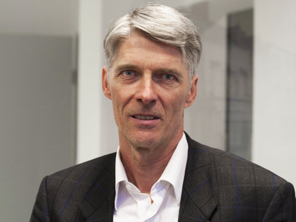 A man with gray hair is wearing a white shirt collar and a dark suit.