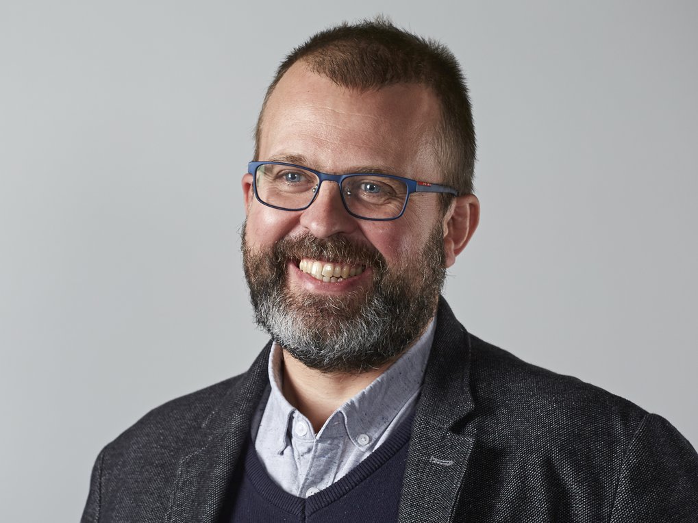 A medium-close-up portrait of a bearded man in business attire—consisting of a shirt, sweater, and blazer—against a neutral background.