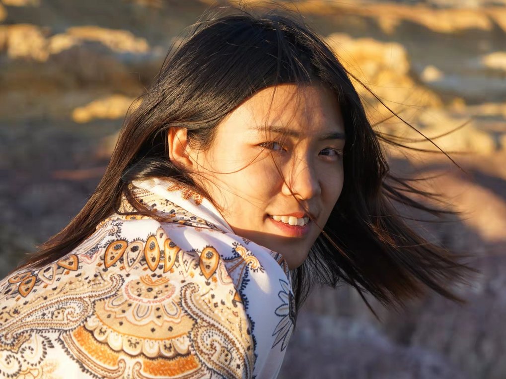 A dark-haired person wearing a colorful scarf with a paisley pattern stands in front of a rocky landscape in the sunlight.