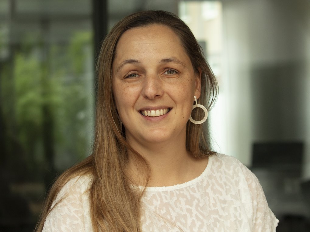A close-up of a woman with long, light brown hair, wearing a white patterned top and large round earrings, against a blurred indoor background.