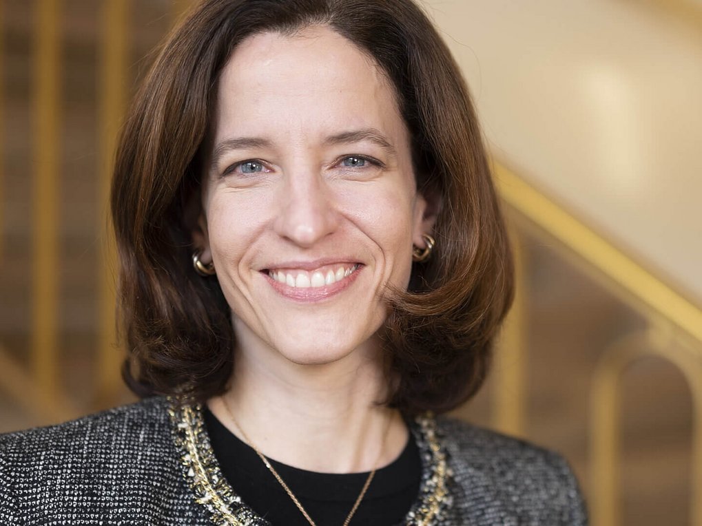 Close-up of a woman with shoulder-length brown hair wearing a gray blazer with a decorative chain; in the background, a staircase with a railing.