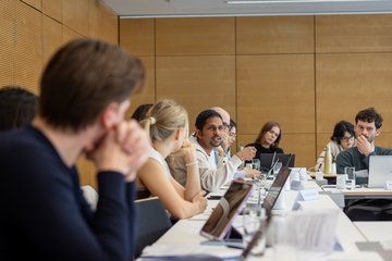 Zu sehen ist eine gruppenhafte Ansammlung von Individuen der Gattung Homo sapiens intellectualis, die in einer modern anmutenden Räumlichkeit an einfachen Tischen sitzend ganz offensichtlich in einem argumentativen Austausch vertieft sind. Allen ist eigen, sich auf dem Tisch einen sogenannten Laptop platziert zu haben: Zweck dürfte sein, sich während der Diskussion Argumente vorbereiten oder bereits vorbereitete Texte vorbringen zu können.