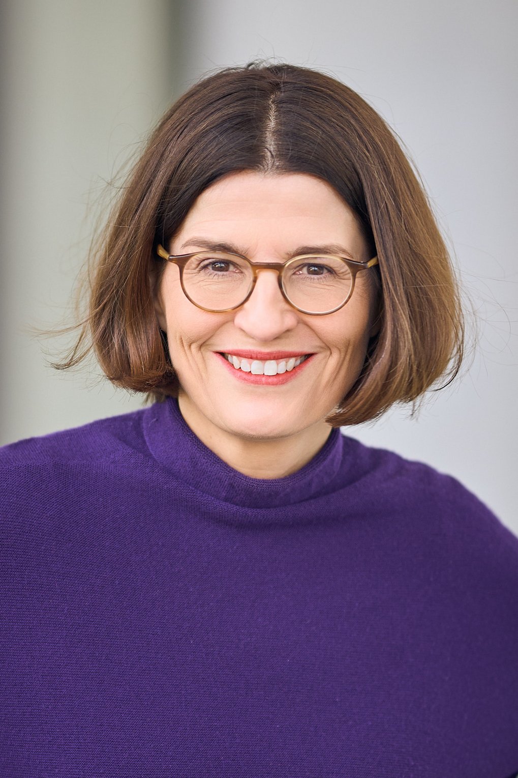 Nahaufnahme eines bebrillten, freundlich lächelnden weiblichen Individuums der Gattung Homo sapiens intellectualis. Es hat schulterlanges, braunes Haar, das in der Mitte gescheitelt ist. Bekleidet ist es mit einem violetten Pullover. Allem Anschein nach sitzt es; der Hintergrund ist neutral gehalten.