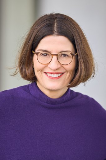 Kia Vahland Nahaufnahme eines bebrillten, freundlich lächelnden weiblichen Individuums der Gattung Homo sapiens intellectualis. Es hat schulterlanges, braunes Haar, das in der Mitte gescheitelt ist. Bekleidet ist es mit einem violetten Pullover. Allem Anschein nach sitzt es; der Hintergrund ist neutral gehalten.