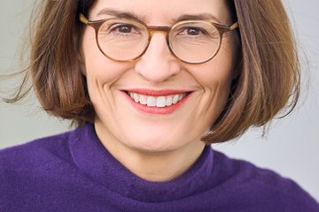 Nahaufnahme eines bebrillten, freundlich lächelnden weiblichen Individuums der Gattung Homo sapiens intellectualis. Es hat schulterlanges, braunes Haar, das in der Mitte gescheitelt ist. Bekleidet ist es mit einem violetten Pullover. Allem Anschein nach sitzt es; der Hintergrund ist neutral gehalten.