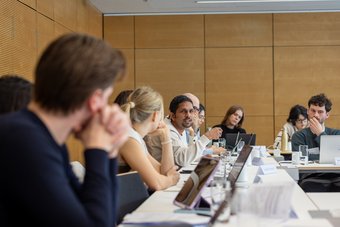 The image shows a group of individuals of the species Homo sapiens intellectualis sitting at simple tables in a modern-looking space, clearly engrossed in a lively discussion. They all have a so-called laptop placed on the table; the purpose is likely to prepare arguments during the discussion or to present texts they have already prepared.