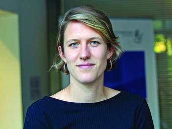 The photo shows a member of the species Homo sapiens intellectualis with short blond hair and hoop earrings, wearing a dark blue sweater, against a blurred background in an interior setting featuring a window and a poster.