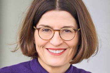 Close-up of a female member of the species Homo sapiens intellectualis, wearing glasses and smiling warmly. She has shoulder-length brown hair parted in the middle. She is wearing a purple sweater. She appears to be sitting; the background is plain.