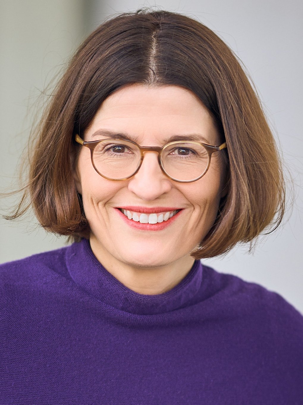 Close-up of a female member of the species Homo sapiens intellectualis, wearing glasses and smiling warmly. She has shoulder-length brown hair parted in the middle. She is wearing a purple sweater. She appears to be sitting; the background is plain.
