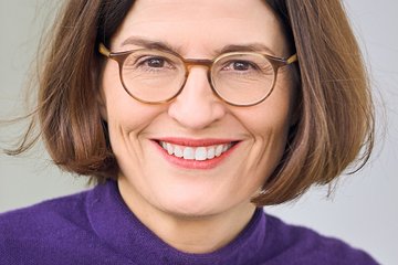 Close-up of a female member of the species Homo sapiens intellectualis, wearing glasses and smiling warmly. She has shoulder-length brown hair parted in the middle. She is wearing a purple sweater. She appears to be sitting; the background is plain.
