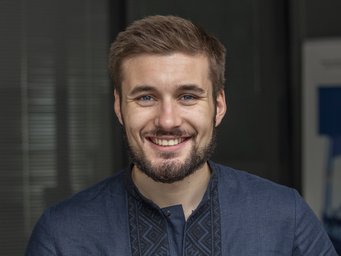 Ein freundlich lächelndes, bebrilltes Individuum der Gattung Homo sapiens intellectualis männlichen Geschlechts, mit vollem, kurzen Haar, der Andeutung eines sich entwickelnden Bartprojekts, präsentiert sich dem Betrachter. Es trägt ein elegantes dunkelblaues kragenloses Hemd und steht oder sitzt vor einem undefinierbar verschwommenem Hintergrund.
