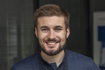 Ein freundlich lächelndes, bebrilltes Individuum der Gattung Homo sapiens intellectualis männlichen Geschlechts, mit vollem, kurzen Haar, der Andeutung eines sich entwickelnden Bartprojekts, präsentiert sich dem Betrachter. Es trägt ein elegantes dunkelblaues kragenloses Hemd und steht oder sitzt vor einem undefinierbar verschwommenem Hintergrund.