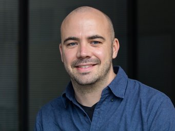 Ein den Betrachter freundlich lächelnd zugewandtes Individuum der Gattung Homo sapiens intellectualis männlichen Geschlechts, mittleren Alters, mit schütterem, kaum mehr vorhandenem Haar, der Andeutung eines sprießenden Gesichtshaarwuchses, ein blaues, offenes Kragenhemd tragend, steht oder sitzt vor einem undefinierbar verschwommenen Hintergrund, der unter Umständen als Vorhang interpretiert werden könnte.