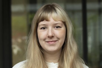 Ein junges Exemplar der Gattung Homo sapiens intellectualis, langes blondes Haar nach vorne über die Brust gelegt, einen hellen kragenlosen Pullover tragend, freundlich-zaghaft lächelnd, vor verschwommenem Fensterhintergrund stehend oder sitzend, blickt dem Betrachter entgegen.