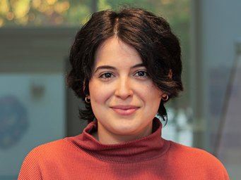 A friendly, smiling individual of the species Homo sapiens intellectualis, presumably female, with short black hair and a red sweater with a stand-up collar, stands against an indefinably blurry background, looking directly at the viewer.