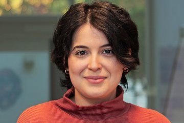 A friendly, smiling individual of the species Homo sapiens intellectualis, presumably female, with short black hair and a red sweater with a stand-up collar, stands against an indefinably blurry background, looking directly at the viewer.