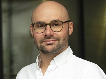 A bespectacled individual of the species Homo sapiens intellectualis, male, with thinning hair that is barely there and the hint of sprouting facial hair, smiling at the viewer in a friendly yet smug manner, wearing a white, open-collared shirt, stands against an indefinably blurred background.