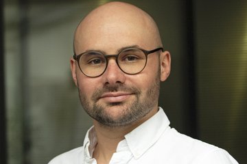 A bespectacled individual of the species Homo sapiens intellectualis, male, with thinning hair that is barely there and the hint of sprouting facial hair, smiling at the viewer in a friendly yet smug manner, wearing a white, open-collared shirt, stands against an indefinably blurred background.