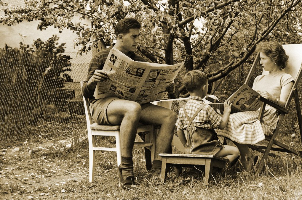 Vater mit Zeitung, Mutter mit Kinderbuch, dazwischen der Sohn: Frühling im Garten, 50er Jahre