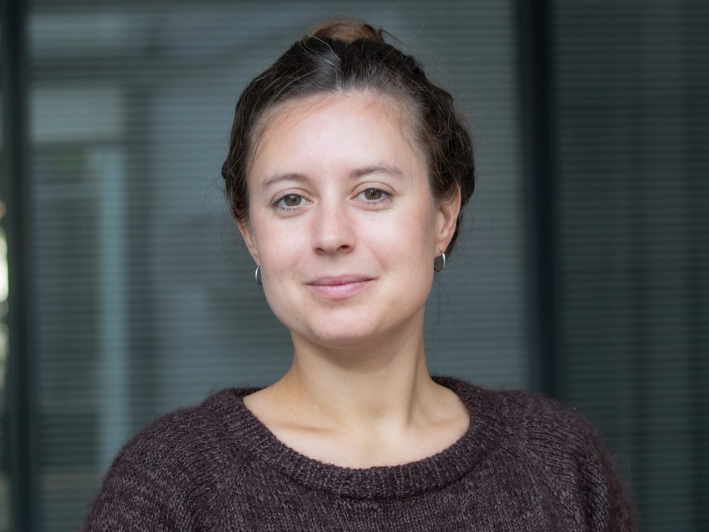 Nahaufnahme einer lächelnden Frau mit zurückgebundenem Haar und braunem Strickpullover vor einem unscharfen, grauen Bürohintergrund.