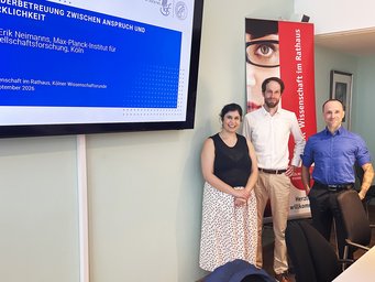 Three people standing in a conference room next to a presentation screen and a banner.
