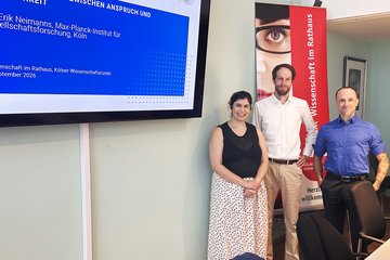 Drei Personen posieren in einem Konferenzraum vor einem Bildschirm mit Präsentationsfolie und einem Banner mit der Aufschrift „Wissenschaft im Rathaus“.