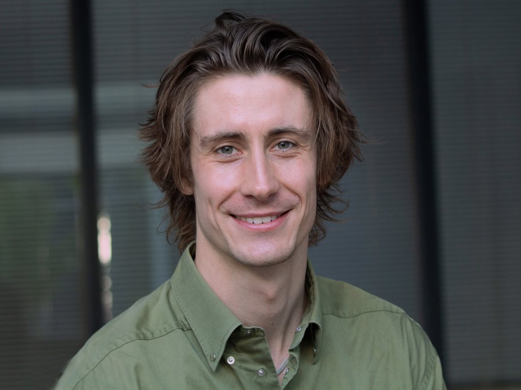 Junger Mann mit mittellangem braunem Haar und grünem Hemd lächelt in die Kamera, unscharfer Hintergrund mit vertikalen Linien.