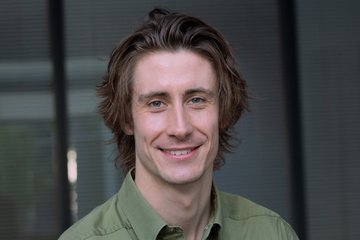 Junger Mann mit mittellangem braunem Haar und grünem Hemd lächelt in die Kamera, unscharfer Hintergrund mit vertikalen Linien.